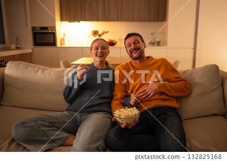 Happy intrigued couple watching movie with popcorn at home cozy together enjoying shared tradition Happy intrigued couple watching movie with popcorn at home cozy together enjoying shared tradition 132825168