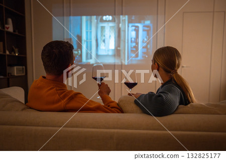 Couple enjoying romantic evening with wine and movie projected on screen at home on sofa rearview 132825177