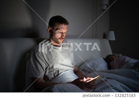 Stressed man awake at night using smartphone during sleepless hours in dark bedroom with wife Stressed man awake at night using smartphone during sleepless hours in dark bedroom with wife 132825183