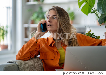 Stunned middle-aged woman receiving sudden shocking news via smartphone while seated at laptop  132825227