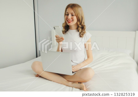 Happy young woman working with laptop using phone sitting on white bed in bedroom at home Happy young woman working with laptop using phone sitting on white bed in bedroom at home 132825319