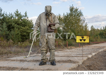 A military man in chemical protection and a gas mask measures the radiation level with a dosimeter, a radiation hazard sign, a concept: radiation leakage, the threat of infection. A military man in chemical protection and a gas mask measures the radiation level with a dosimeter, a radiation hazard sign, a concept: radiation leakage, the threat of infection. 132825349