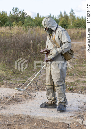 A military man in chemical protection and a gas mask measures the radiation level with a dosimeter in nature, smoke from a fire. Concept: radiation leakage, radioactive hazard. 132825427