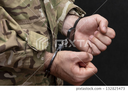 A soldier in a military camouflage uniform is handcuffed on a black background. 132825473