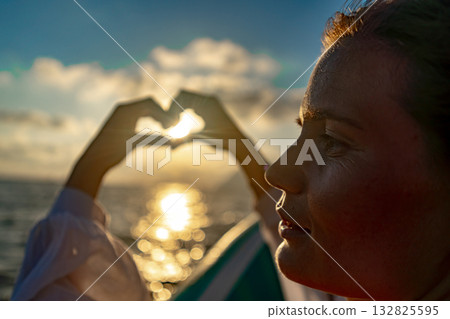 Hands, heart, sunset. Woman forming heart shape with hands, framing golden light over tranquil ocean. Hands, heart, sunset. Woman forming heart shape with hands, framing golden light over tranquil ocean. 132825595