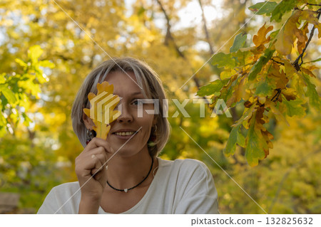 Woman autumn leaf holding yellow oak leaf covering one eye smiling happily in park Woman autumn leaf holding yellow oak leaf covering one eye smiling happily in park 132825632
