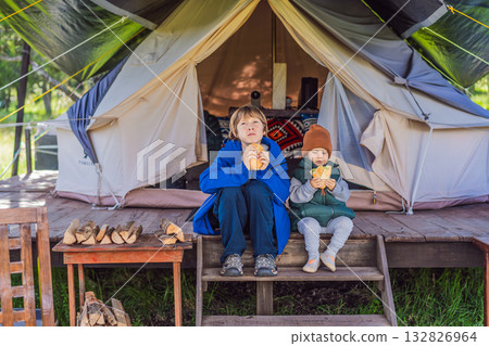 Toddler and his teenage brother tourists in warm vests and hats eating buns while sitting on glamping steps, enjoying family travel, outdoor adventure, and bonding Toddler and his teenage brother tourists in warm vests and hats eating buns while sitting on glamping steps, enjoying family travel, outdoor adventure, and bonding 132826964