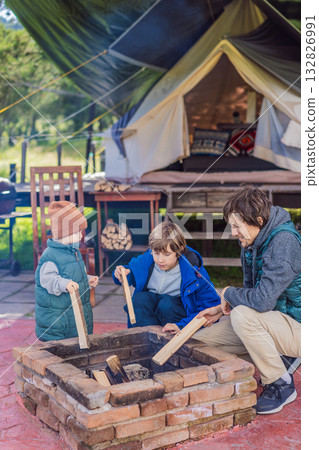 Father and his two sons making a campfire in front of a glamping site, enjoying family bonding, outdoor adventure, and nature exploration 132826991