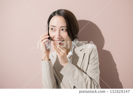 Smiling businesswoman in a suit talking on a smartphone on a pink background, career woman, secret conversation Smiling businesswoman in a suit talking on a smartphone on a pink background, career woman, secret conversation 132828147