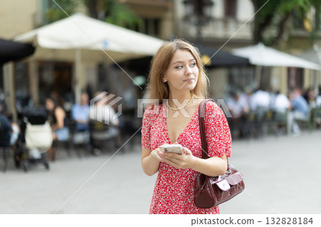 Girl in summer dress walks along street of the old town with mobile phone in her hands 132828184