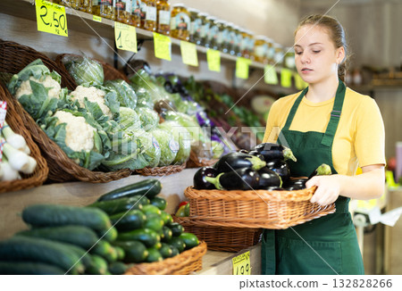 Girl seller in uniform working in supermarket at her first job, checking organic eggplants Girl seller in uniform working in supermarket at her first job, checking organic eggplants 132828266