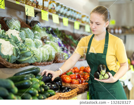 Teenage girl seller puts eggplants in vegetable shop 132828327
