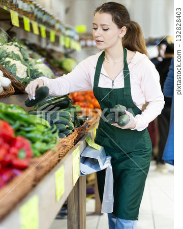 Young woman seller puts cucumbers in vegetable shop 132828403