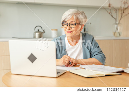 Online education courses webinar concept. Middle aged senior woman using laptop computer writing notes. Focused mature old woman enjoying studying online from home written records doing online work Online education courses webinar concept. Middle aged senior woman using laptop computer writing notes. Focused mature old woman enjoying studying online from home written records doing online work 132834527