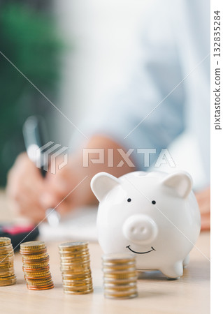 Concept of saving money and investment planning. A businesswoman drops coin into white piggy bank on office desk with documents, symbolizing financial security, budget management and future wealth. Concept of saving money and investment planning. A businesswoman drops coin into white piggy bank on office desk with documents, symbolizing financial security, budget management and future wealth. 132835284