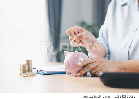 Concept of saving money and financial security. Pink piggy bank surrounded by coins on office desk with businesswoman in background, symbolizing investment, wealth growth and budget management. 132835523