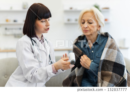 A female doctor in a white coat checks an elderly woman's temperature with a digital thermometer. The patient is wrapped in a blanket. A female doctor in a white coat checks an elderly woman's temperature with a digital thermometer. The patient is wrapped in a blanket. 132837777