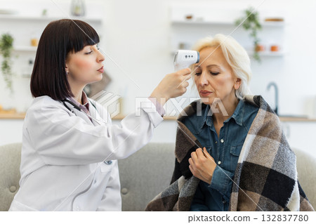 A female doctor uses a contactless thermometer to check the temperature of an elderly woman. The patient is wrapped in a blanket. A female doctor uses a contactless thermometer to check the temperature of an elderly woman. The patient is wrapped in a blanket. 132837780