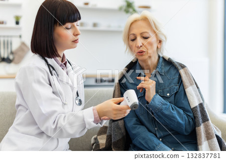 A female doctor checks the temperature of an elderly woman using a digital thermometer during a home visit. The patient is wrapped in a blanket. A female doctor checks the temperature of an elderly woman using a digital thermometer during a home visit. The patient is wrapped in a blanket. 132837781