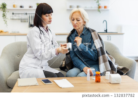 A female doctor checks the temperature of an older woman who is sitting on a couch, possibly feeling unwell. Medications are on the table. A female doctor checks the temperature of an older woman who is sitting on a couch, possibly feeling unwell. Medications are on the table. 132837782