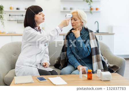 A doctor uses a non-contact thermometer to check an elderly woman's temperature while she is sitting on a couch at home. A doctor uses a non-contact thermometer to check an elderly woman's temperature while she is sitting on a couch at home. 132837783