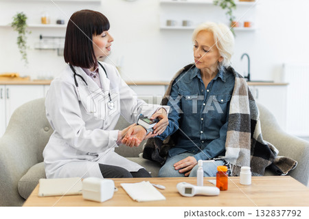 A female doctor in a white coat measures the blood pressure of an elderly woman sitting on a sofa, providing at-home healthcare. 132837792