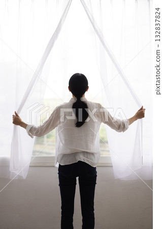 一位女士背對著鏡頭，拉開窗簾迎接晨光。 132837824