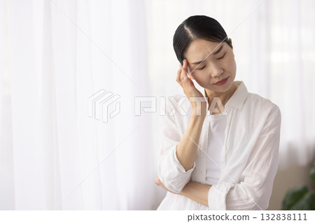 A middle-aged woman furrows her brow and places her hand on her temple by the window A middle-aged woman furrows her brow and places her hand on her temple by the window 132838111