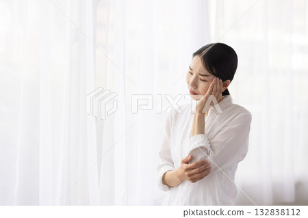 A middle-aged woman furrows her brow and places her hand on her temple by the window A middle-aged woman furrows her brow and places her hand on her temple by the window 132838112