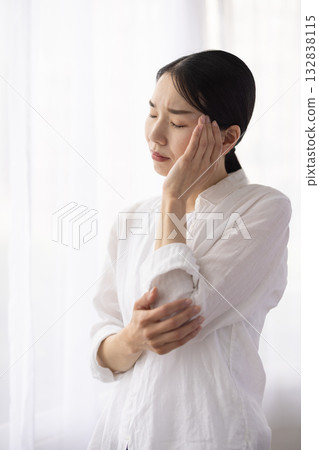 A middle-aged woman furrows her brow and places her hand on her temple by the window A middle-aged woman furrows her brow and places her hand on her temple by the window 132838115