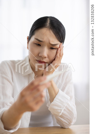 A middle-aged woman looks at a thermometer, puts her hand on her head, and frowns A middle-aged woman looks at a thermometer, puts her hand on her head, and frowns 132838206