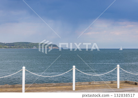 The coast of the Baltic Sea in northern Poland. A serene seascape featuring calm blue waters, a distant sailboat, and a lush green shoreline, framed by a wooden dock and white posts. 132838517