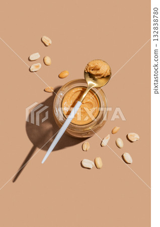 Peanut butter in a glass jar on a beige background with a spoon, scattered nuts and a shadow. 132838780