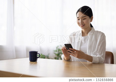 一位微笑的中年婦女正在桌邊操作智慧型手機。 132838866