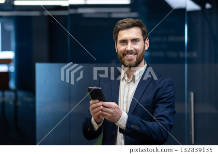 Portrait of a smiling young businessman in a suit standing in an office, holding a phone, looking at the camera. 132839135