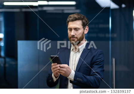A successful young man in a suit is standing inside the office and using a mobile phone with a smile. 132839137