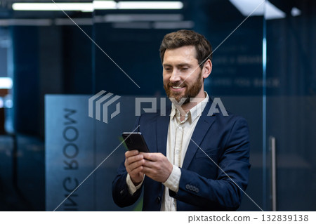 A close-up photo of a young smiling businessman in a suit standing in the office and using a mobile phone. A close-up photo of a young smiling businessman in a suit standing in the office and using a mobile phone. 132839138
