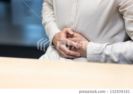 A close-up photo of the hands of a young man in a light shirt sitting at a desk in the office and holding his stomach. A close-up photo of the hands of a young man in a light shirt sitting at a desk in the office and holding his stomach. 132839181