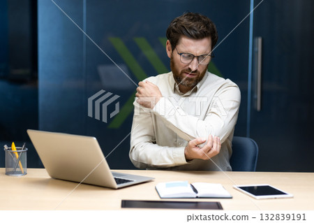 A young man, grimacing in pain, sits at a desk in an office and holds his elbow with his hand, feeling severe pain. A young man, grimacing in pain, sits at a desk in an office and holds his elbow with his hand, feeling severe pain. 132839191