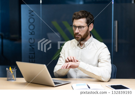 Worried young man sitting in the office, working on a laptop and scratching his hand from allergies. Worried young man sitting in the office, working on a laptop and scratching his hand from allergies. 132839193
