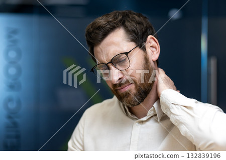 Close-up photo of a tired young man working in an office, rubbing his neck with his hand, feeling pain and tension. 132839196