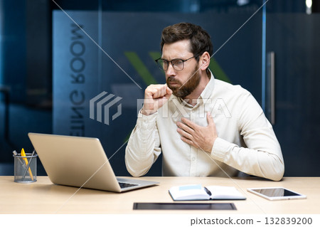 A young male office worker sits at a desk in the office, holds his hand to his chest and experiences severe coughing fits. A young male office worker sits at a desk in the office, holds his hand to his chest and experiences severe coughing fits. 132839200