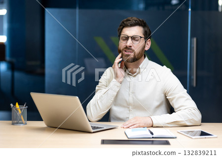 A young businessman suffering from pain sits in the office at a desk and holds his cheek with his hand, feeling severe pain and discomfort. 132839211