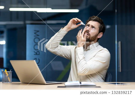 A young male office worker sits at a desk in the office and puts medical drops in his eyes. A young male office worker sits at a desk in the office and puts medical drops in his eyes. 132839217
