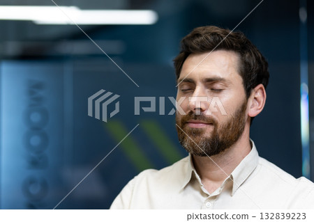 Close-up photo of a young man in a shirt and with a beard, closing his eyes, breathing deeply and relaxing during working hours in the office. 132839223