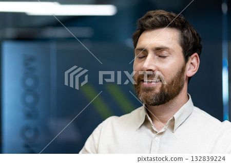 Close-up photo of a calm young man in a shirt and with a beard who is in the office, sitting at his workplace, closing his eyes and resting. 132839224