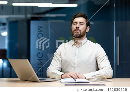 A young man sits in the office at his workplace, his eyes closed and resting. A young man sits in the office at his workplace, his eyes closed and resting. 132839226