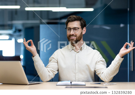 A relaxed young man works in the office, sits at his desk in the lotus position during a break and meditates, looking at the laptop screen. A relaxed young man works in the office, sits at his desk in the lotus position during a break and meditates, looking at the laptop screen. 132839280