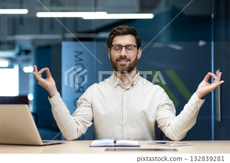 A young office worker is resting at his desk, sitting in the lotus position with his eyes closed and meditating. A young office worker is resting at his desk, sitting in the lotus position with his eyes closed and meditating. 132839281