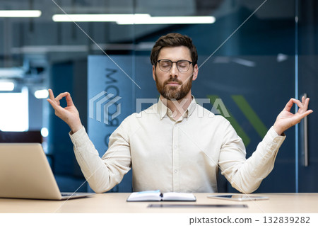 Young male businessman resting and taking a break in the office, sitting at his desk in the lotus position, closing his eyes and meditating. Young male businessman resting and taking a break in the office, sitting at his desk in the lotus position, closing his eyes and meditating. 132839282